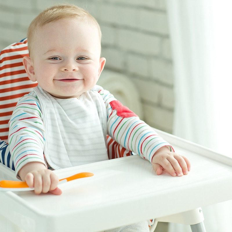 Toddler in a high chair holding a spoon and smiling